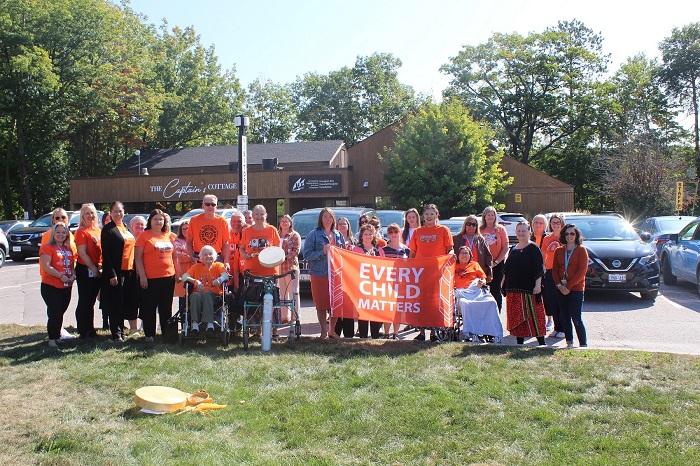 Grand groupe de personnes de tous les âges qui portent des chandails orange « Chaque enfant compte » et tiennent une bannière correspondante.