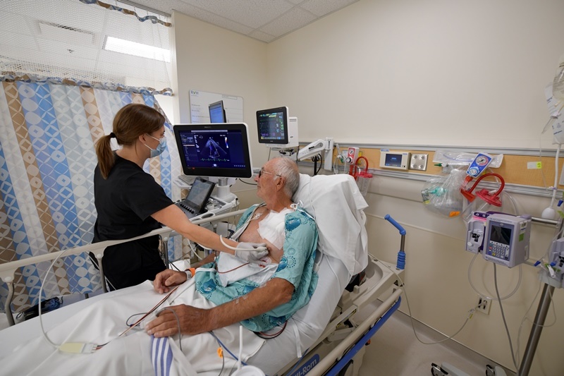 A patient in a hospital bed with tubes strapped to him. An attendant is looking at a screen while performing an echocardiography.