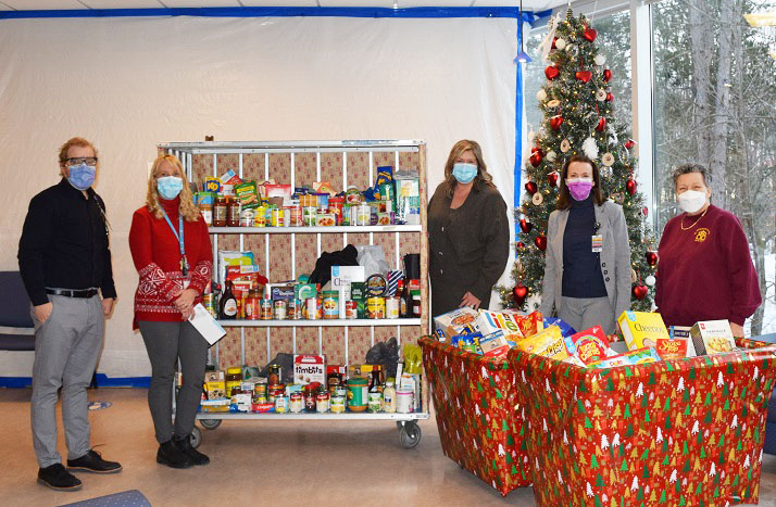 Four people wearing masks surrounded by shelving and 2 large bins full of donated food for the food drive.