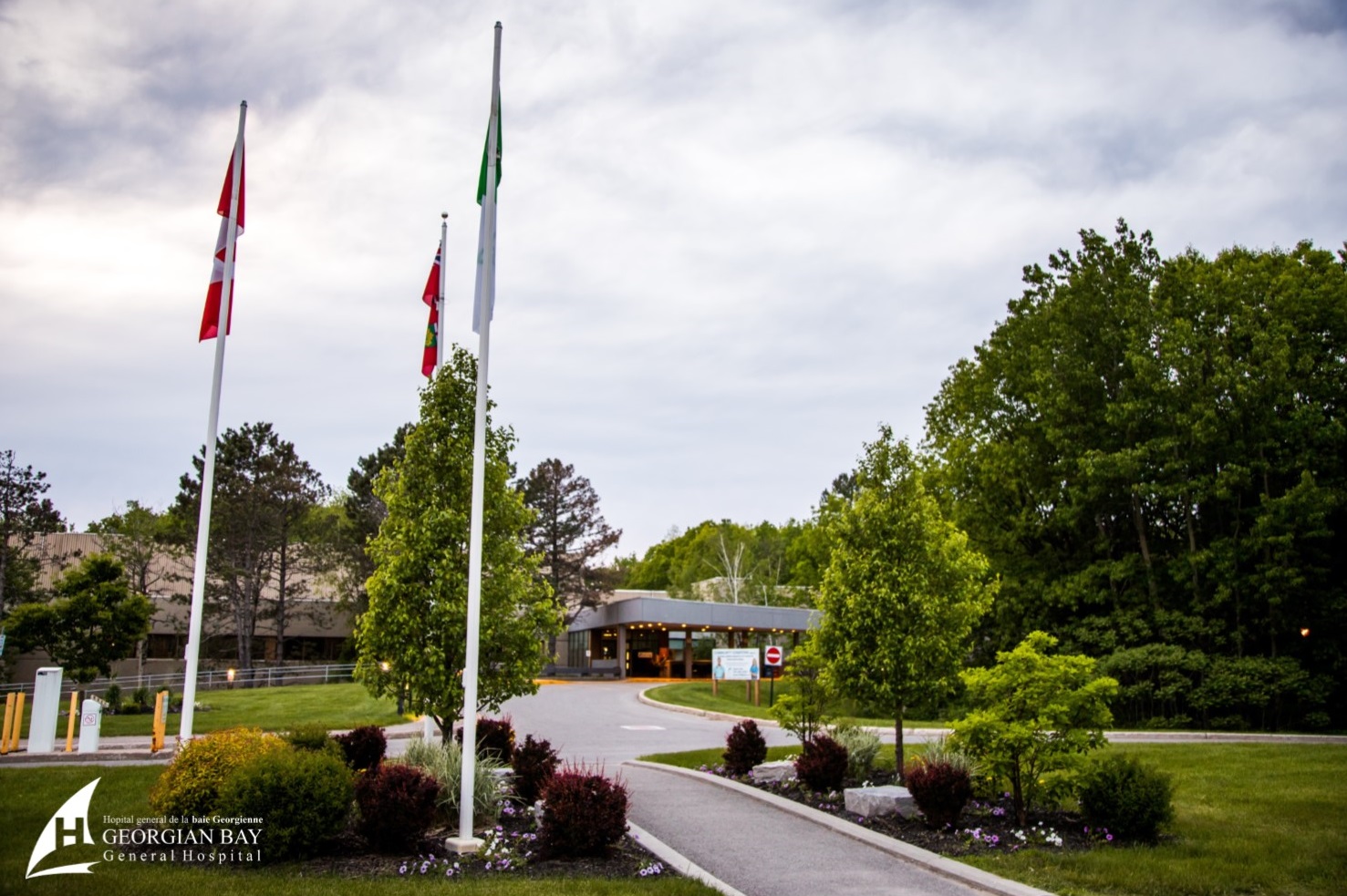 The main entrance of the hospital showing the walkway, 2 flag posts, and a nice garden with green trees and plants in bloom.