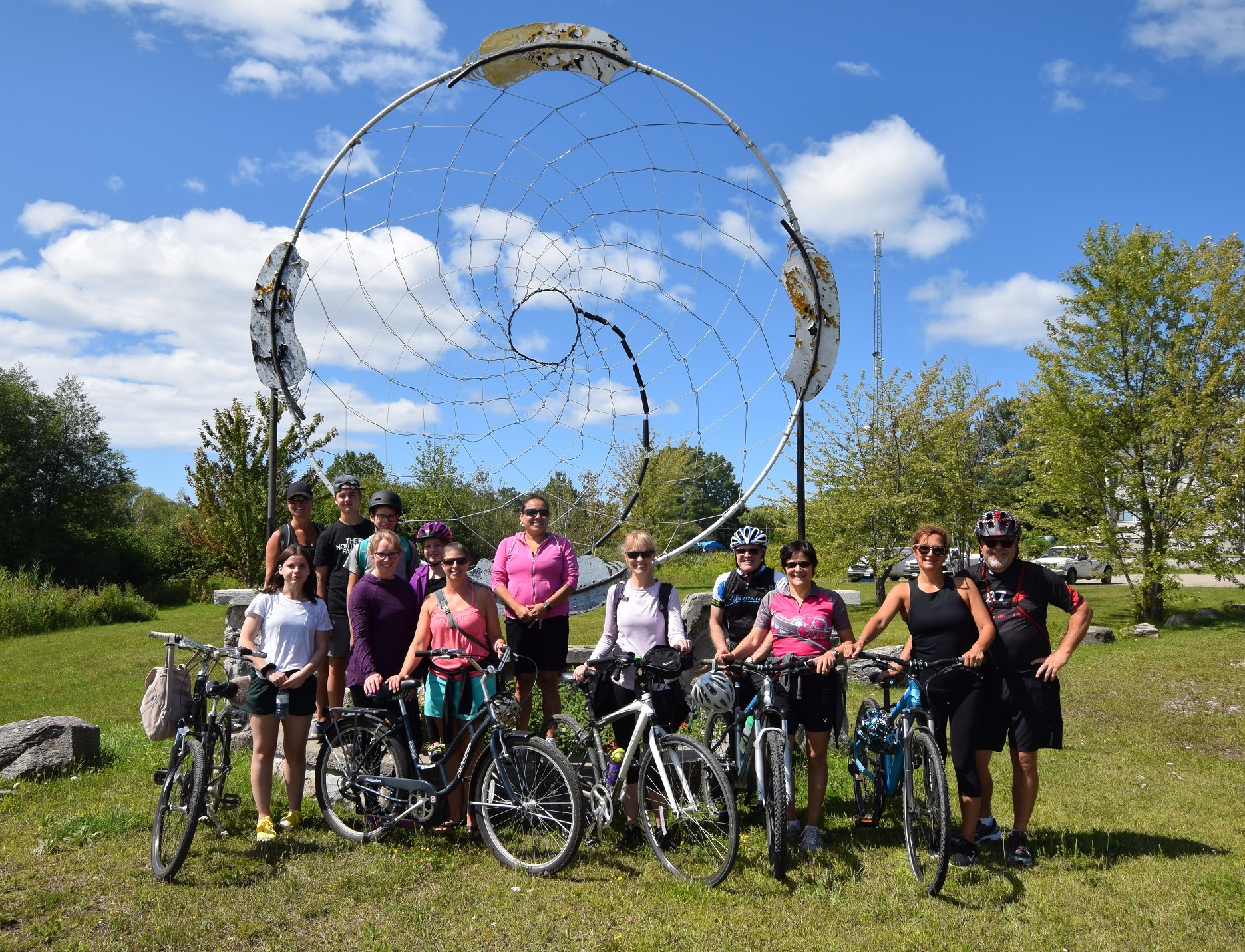 Twelve people of all ages with bicycles and helmets pose in front of a very large metals dream catcher art piece.