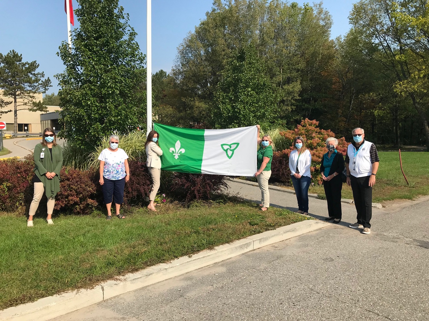 Six women and one man stand distanced wearing masks. Two of the people hold up the green and white Franco-Ontario flag.