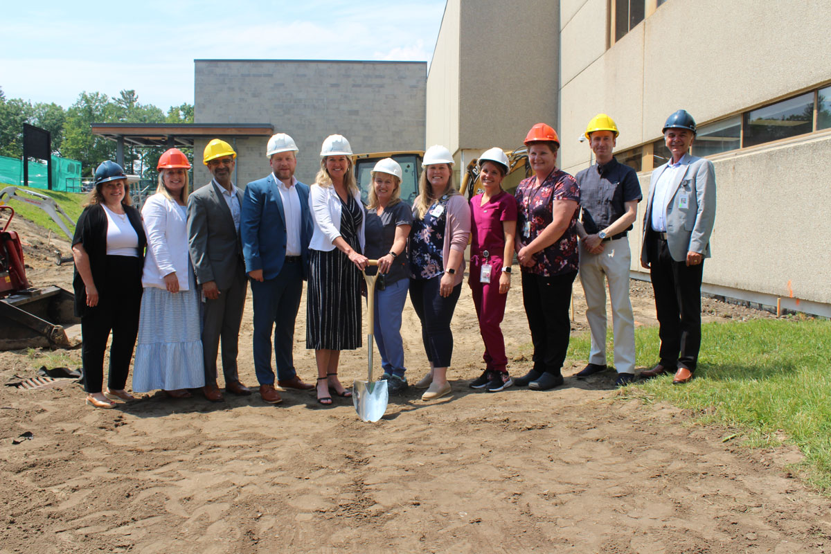 Eleven people wearing coloured hard hats standing on a dirt ground. There is a shovel held by two of the people showing that the dirt is ready to dig.