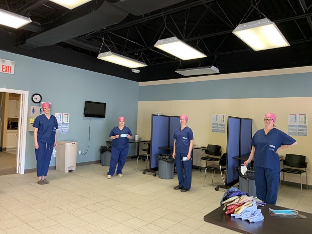 Four women in scrubs and pink bandanas in a room with 3 desks set up with dividing walls between each.