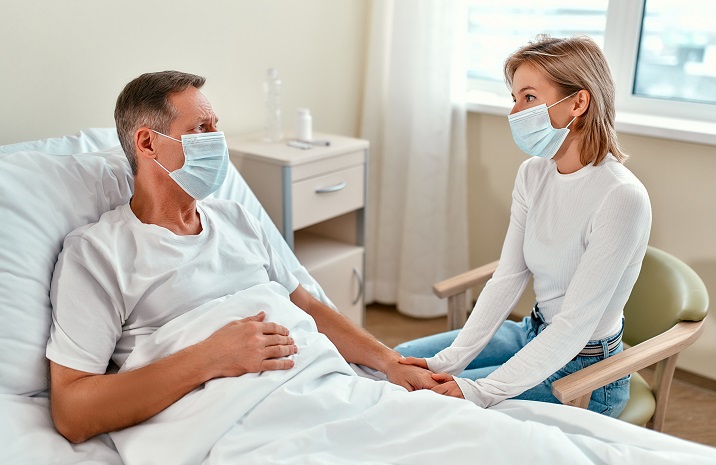 Woman visiting a man in a hospital bed. Both are wearing masks.