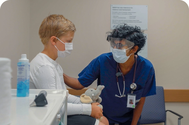 A young blond boy sits on a table with a stuffed elephant while a doctor in scrubs speaks to him.