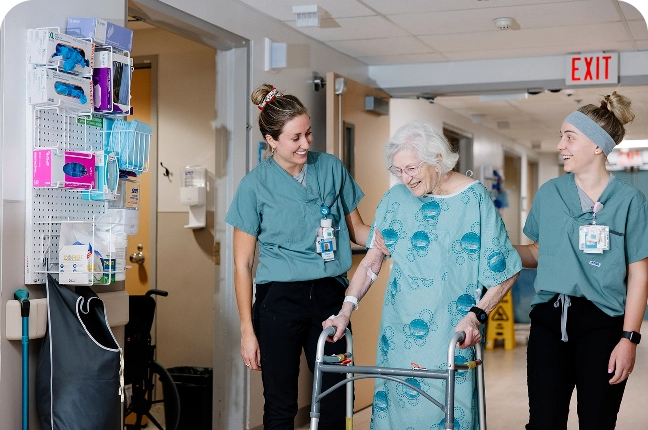 A white haired elderly woman wearing a hospital gown walks using a walker while 2 hospital workers assist her.